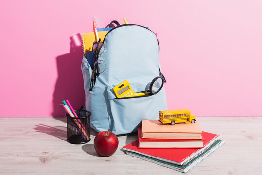 full backpack with school stationery near toy school bus on books, ripe apple and pen holder on pink
