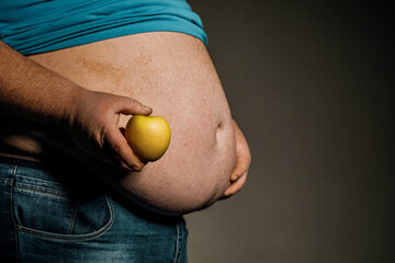 The fat man holds a Apple in front of his bare stomach. The concept of diet and healthy eating