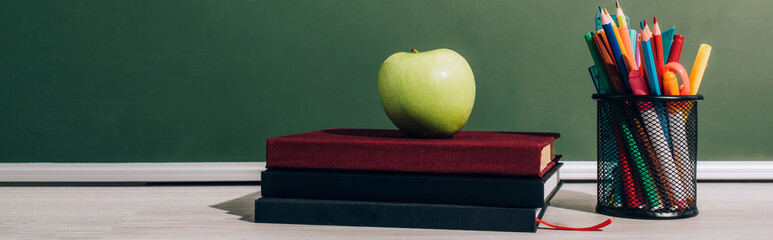 horizontal image of ripe apple on books near pen holder with color pencils near green chalkboard
