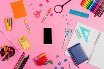 Top view of smartphone with blank screen surrounded by school supplies on pink