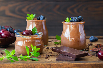 Portion jars with chocolate mousse or pudding decorated with sweet cherry, blueberry and mint and pieces of chocolate. Selective focus. Rustic style. Dark background.