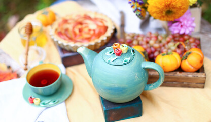 Autumn picnic. Tea party with beautiful kettle, cups at wooden table in garden. Harvest festival. Honey with stick, spoon, apple pie, persimmons, grapes, maple leaf, flowers, yellow linen tablecloth