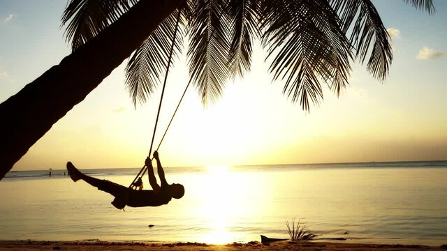 Silhouettes of a guy riding on a swing attached to a palm tree on the ocean at sunset