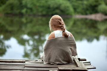 The girl sits on the pier and looks at the lake. Wraps up in a shawl (scarf) - a good day. The...