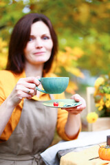 Autumn picnic. Woman in yellow dress and linen apron drinks tea from cup at wooden table in garden. Beautiful kettle, tablecloth, honey with spoon, apple pie, harvest, persimmon, grapes, maple leaf