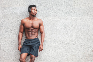 african american athletic man with athletic body stands outdoors against a wall and listens to music, a serious macho without a shirt