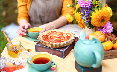 Autumn picnic. Woman in yellow dress and linen apron drinks tea from cup at wooden table in garden. Beautiful kettle, tablecloth, honey with spoon, apple pie, harvest, persimmon, grapes, maple leaf