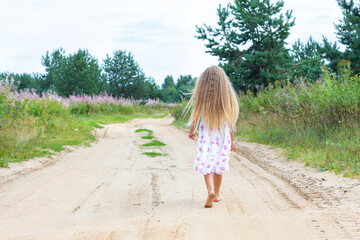 A girl with long blond curly hair walks along a rural road. Full-length portrait from the back. A walk in the fresh air in solitude, reuniting with nature. Beautiful summer landscape.