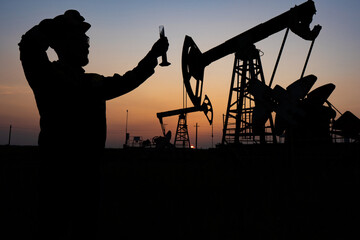Silhouette of a chemical engineer taking a sample of crude oil against the background of oil pumps.