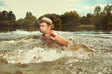 Funny man in stylish retro swimsuit and straw hat swimming in river