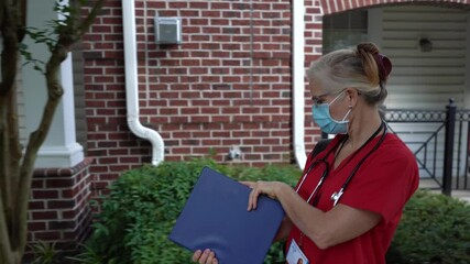 Closeup of side view of home health care nurse wearing a medical face mask walking holding a notebook and wearing a stethoscope and walking on a sidewalk to client’s apartment.