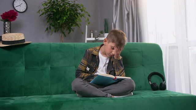 Handsome concentrated teen boy is tired to read uninteresting book,sitting on green soft couch at home