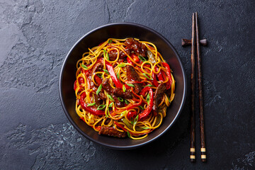Stir fry noodles with vegetables and beef in black bowl. Slate background. Top view.