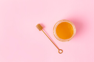 Honey in a glass jar and a golden honey spoon nearby on a pink background. View from above.