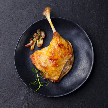 Duck leg confit with mushroom sauce on black plate. Slate background. Top view.