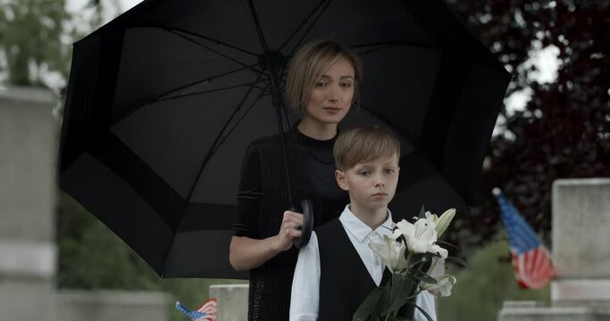 Upset mom embracing her son while standing near gravestone with american flag under umbrella. Widow and young boy with flowers honoring soldier at cemetery. Concept of memorial day