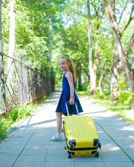 A smiling little girl in a blue dress is walking along the street with a yellow suitcase. A child is going on a summer vacation trip.