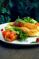 Variation of smorrebrod on rye bread with omelette, cucumbers and salmon in a white plate on a wooden background