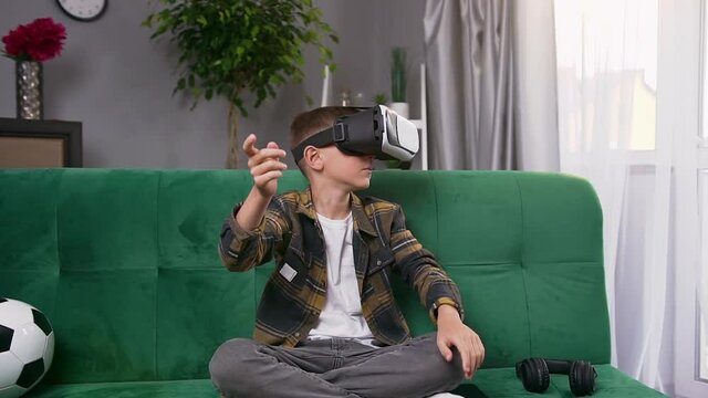 Likable serious modern teen boy sitting on soft green couch and using special futuristic 3d glasses to play interesting video game