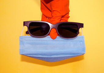 Hand in protective glove holding face mask and sunglasses. Global quarantine and social distancing during summer time. Accessories for infection prevention.