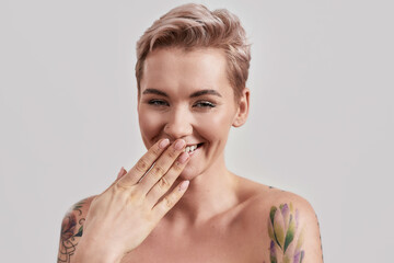 Funny. Portrait of a young attractive half naked tattooed woman with perfect skin laughing at camera, covering mouth with her hand isolated over light background