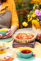 Autumn picnic. Woman in yellow dress and linen apron drinks tea from cup at wooden table in garden. Beautiful kettle, tablecloth, honey with spoon, apple pie, harvest, persimmon, grapes, maple leaf