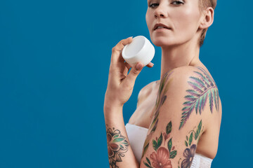 Cropped portrait of beautiful half naked tattooed woman advertising, holding white plastic jar of...