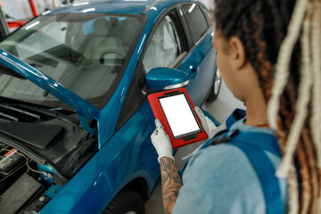 Right repair. Young african american woman, professional female mechanic looking at the blank screen of tablet pc, standing near car with open hood at auto repair shop
