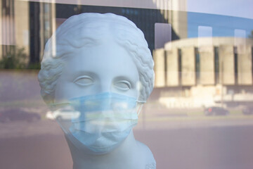  a man, woman plaster bust in a protective medical mask behind a plate-glass window with reflection...