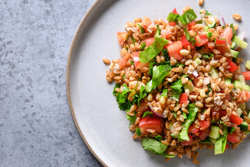 Salad of whole grain cereal spelt with vegetables, tomato, cucumber, greens on grey stone table...