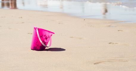 Plastic pink toy bucket left alone on the beach sand near water, blurred ocean on background - concept of vacation safety, drowning danger, also pollution by messy tourists