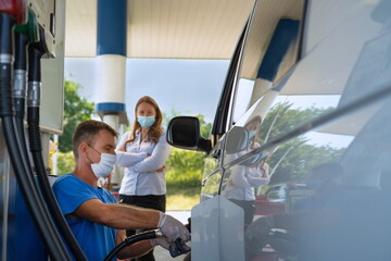 A worker with a mask on his face at the gas station refuel the van while angry customer waits