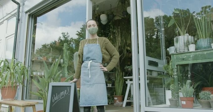 Small business entrepreneur female opening plant shop with welcome sign wearing face mask during coronavirus pandemic and smiling to camera