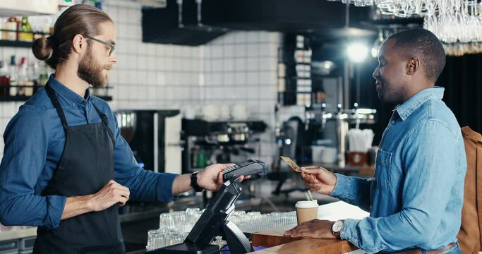 Joyful African American man client in cafe paying with credit card. Mixed-races. Caucasian bartender giving order and taking money. Multiethnic customer and barrista. Pay for coffee drink with paypass