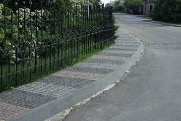 black fence made of iron rods in green vegetation near a stone sidewalk and a gray asphalt road
