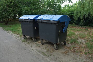 two black large plastic garbage cans closed with blue lids stand on green grass and vegetation in a park by an asphalt gray road