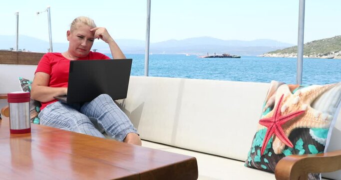 Portrait of a middle aged freelancer woman is sitting on the deck of a yacht with a laptop computer at summer vacation