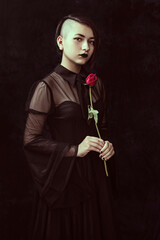 portrait of a goth teenager wearing a black dress with shaved hair on one side holding a red rose