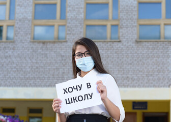 A schoolgirl in a medical mask in the background of the school. A high school student stands near the school. A girl in an mask holds a leaflet with an inscription. Translation: I want to go to school