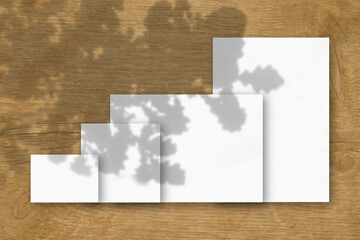 Several horizontal and vertical sheets of white textured paper against a wooden table background. Mockup with an overlay of plant shadows. Natural light casts shadows from the oak leaves