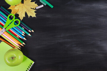 Colorful pencils and crayons, fresh apple, yellow leaf, scissors, green notebook on chalk board...