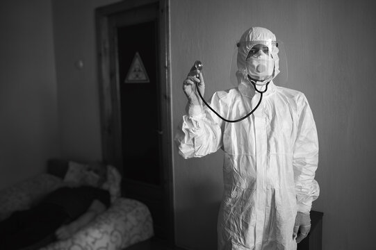 Black And White Image Of Doctor In PPE Suit, Respirator, Gloves, Visiting Patient At Home During Covid-19 Quarantine, Holding Phonendoscope. Biohazard Sign On Door And Patient On Background