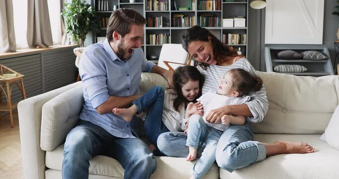 Happy young married couple having fun with cute little daughters on comfortable sofa. Cheerful family spouses parents playing, enjoying spending free weekend time at home with adorable children.