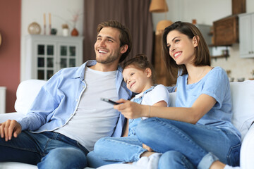 Happy family with child sitting on sofa watching tv, young parents embracing daughter relaxing on couch together.