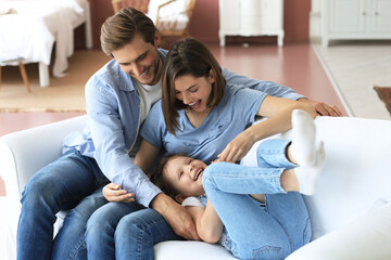 Positive friendly young parents with smiling little daughter sitting on sofa and having fun...