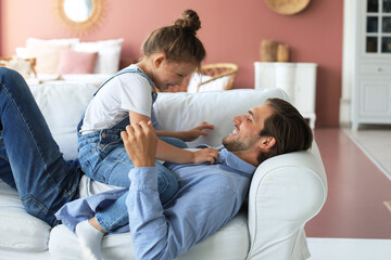 Fototapeta premium Smiling young father lying on couch at living room and play with happy little daughter.