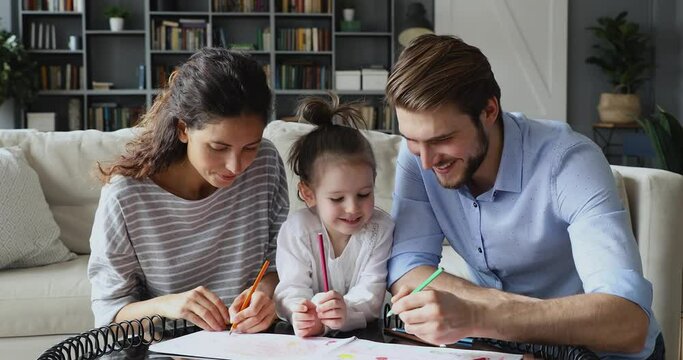Happy caring couple drawing pictures in paper album with adorable little kid daughter in living room. Affectionate young parents helping educating painting small child, involve in creative activity.