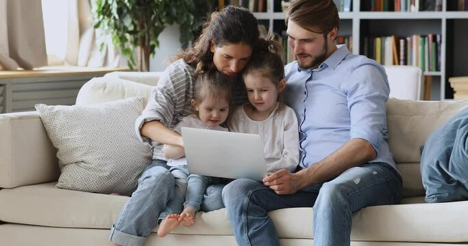 Happy young couple parents showing educational application on computer to little cute children daughters at home, early education concept. Smiling mum and dad watching cartoons with kids on laptop.