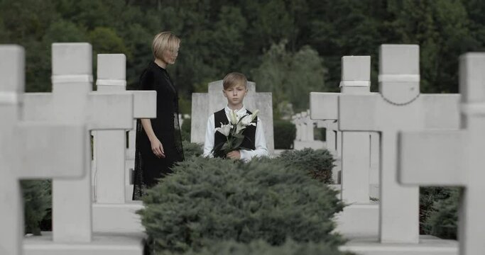 Young kid holding white lily flower at cemetery while his mother coming and embracing him.Teen boy looking to camera while standing in row of stone crosses. Concept of memorial day