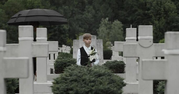 Mother in mourning clothes hugging her young kid with flower at cemetery.Teen boy and sad young widow standing in row of stone crosses under umbrella. Concept of memorial day